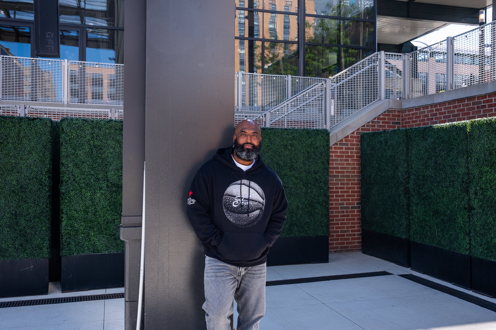 A man wearing a black hoodie with a circular graphic print and light gray jeans, standing outdoors next to a dark gray column in front of green hedges and a modern building.