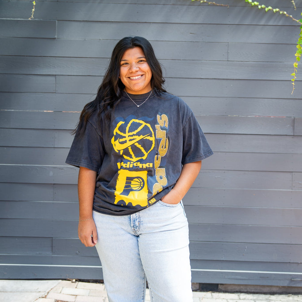 Person wearing a graphic t-shirt standing in front of a gray wall with greenery.
