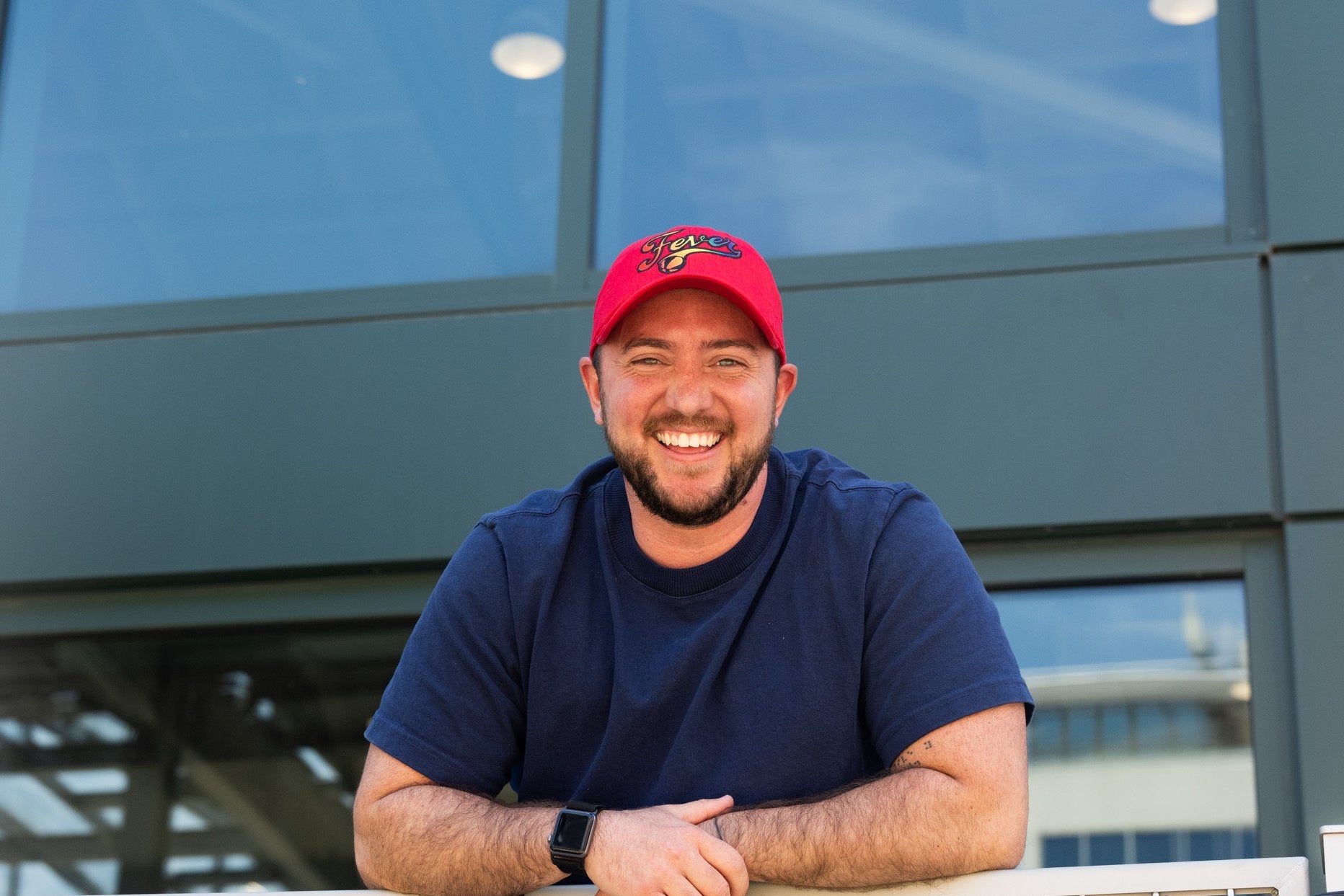 Man wearing a red cap and blue shirt leaning against a railing with a building in the background