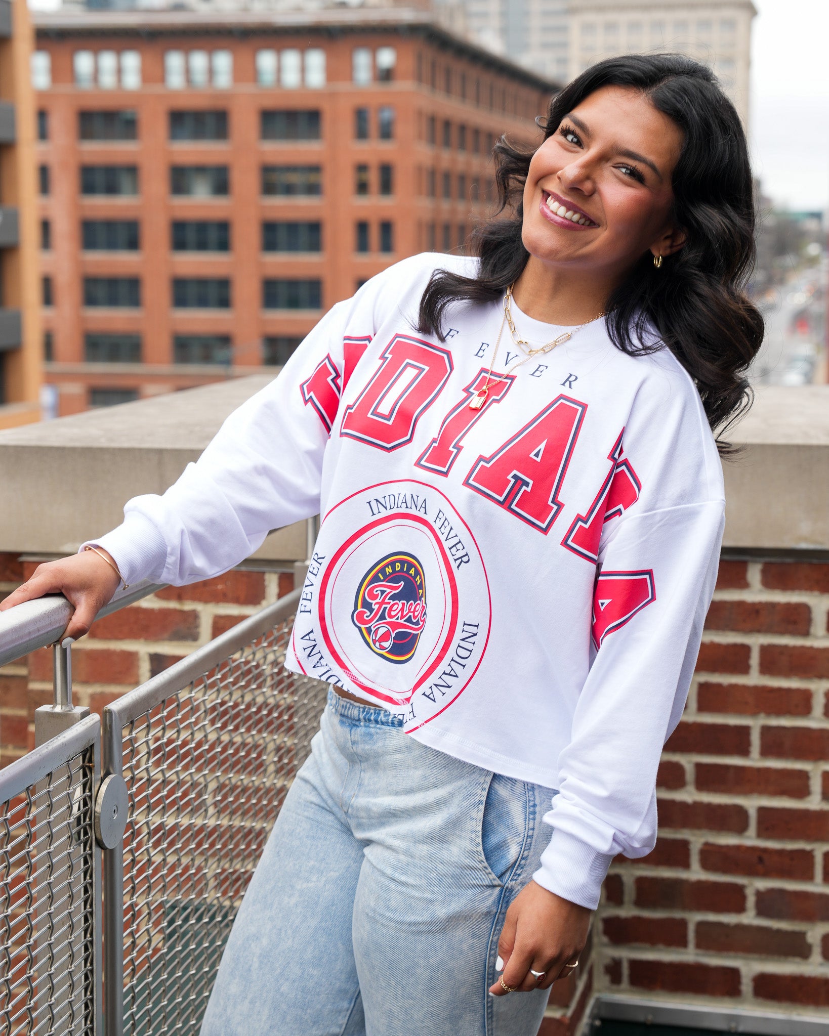 Woman wearing a white sweatshirt with red text and graphics, standing on a rooftop with a cityscape background.