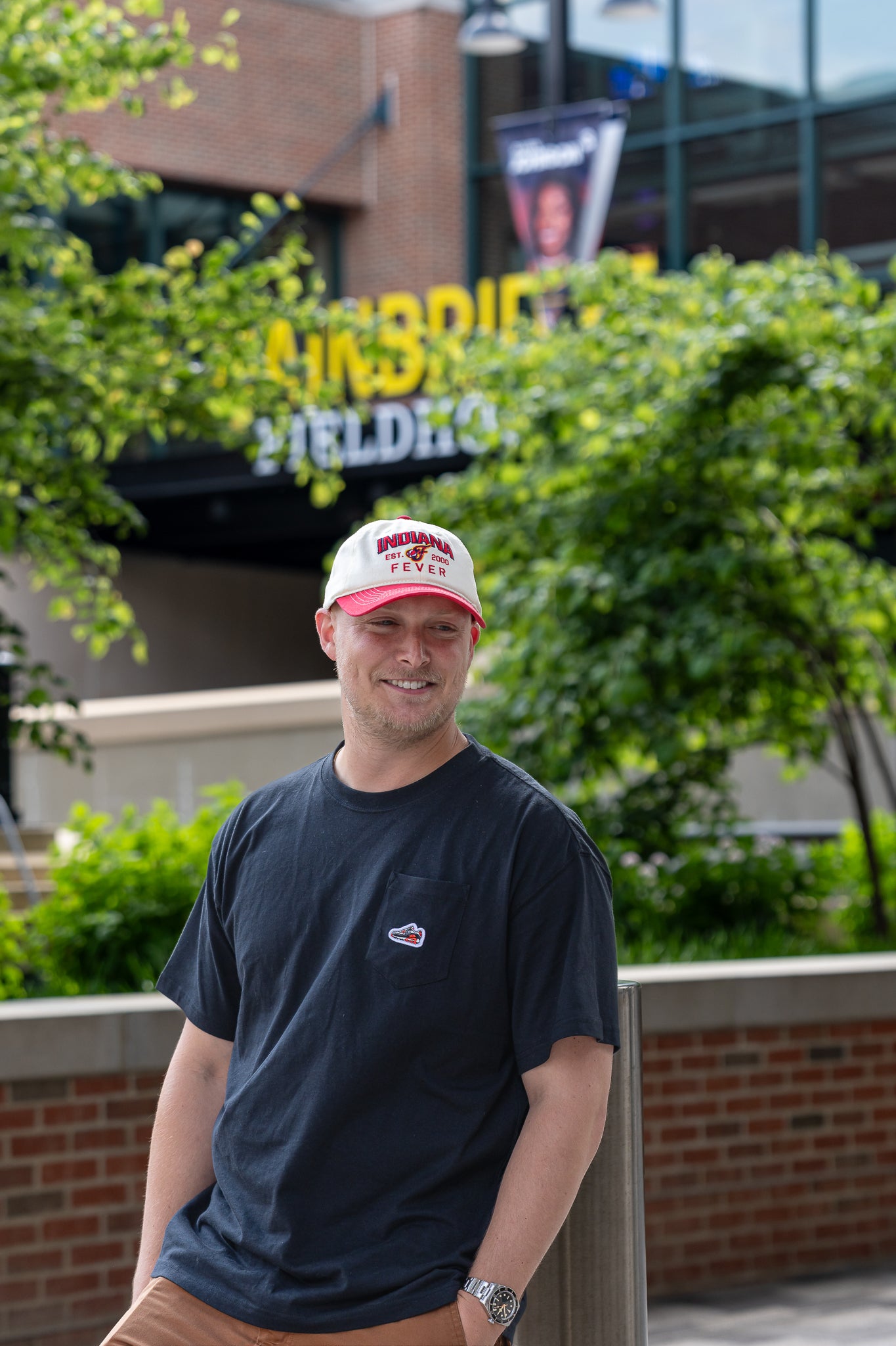 Man wearing a cap and dark shirt standing outdoors with greenery and a building in the background