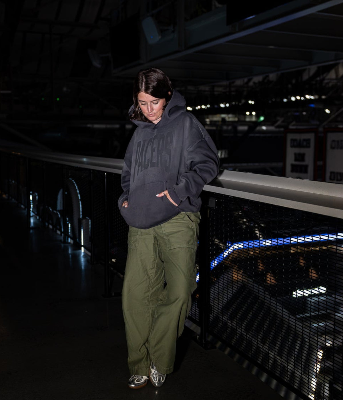 A person standing on a dark indoor walkway or balcony, wearing a dark navy hoodie, olive green pants, and white sneakers. The background is dimly lit with industrial ceiling structures and railing details.