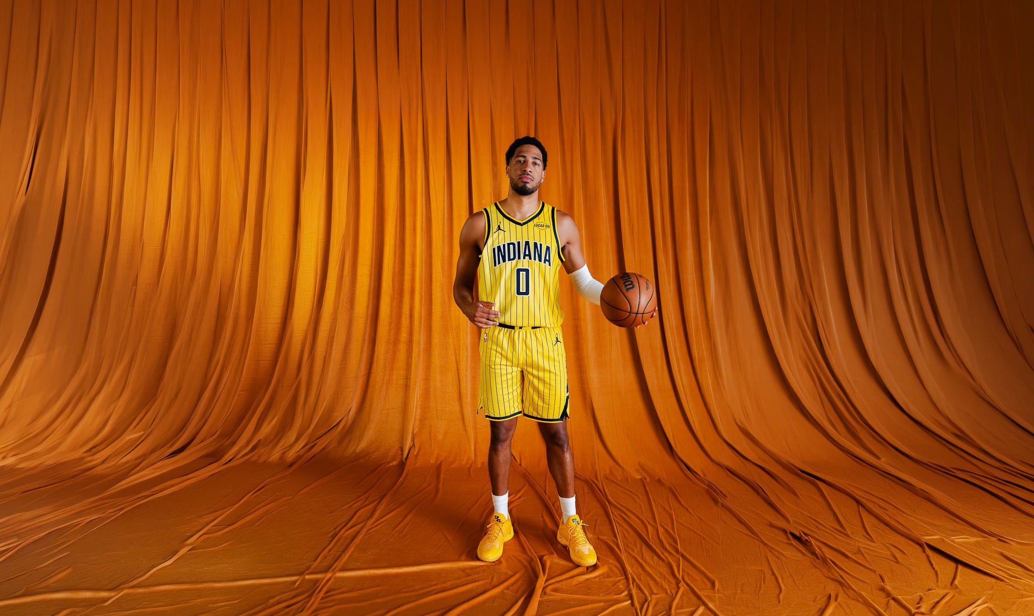 A basketball player stands in a studio wearing a yellow Indiana Pacers jersey and shorts, holding a basketball against a draped orange backdrop.