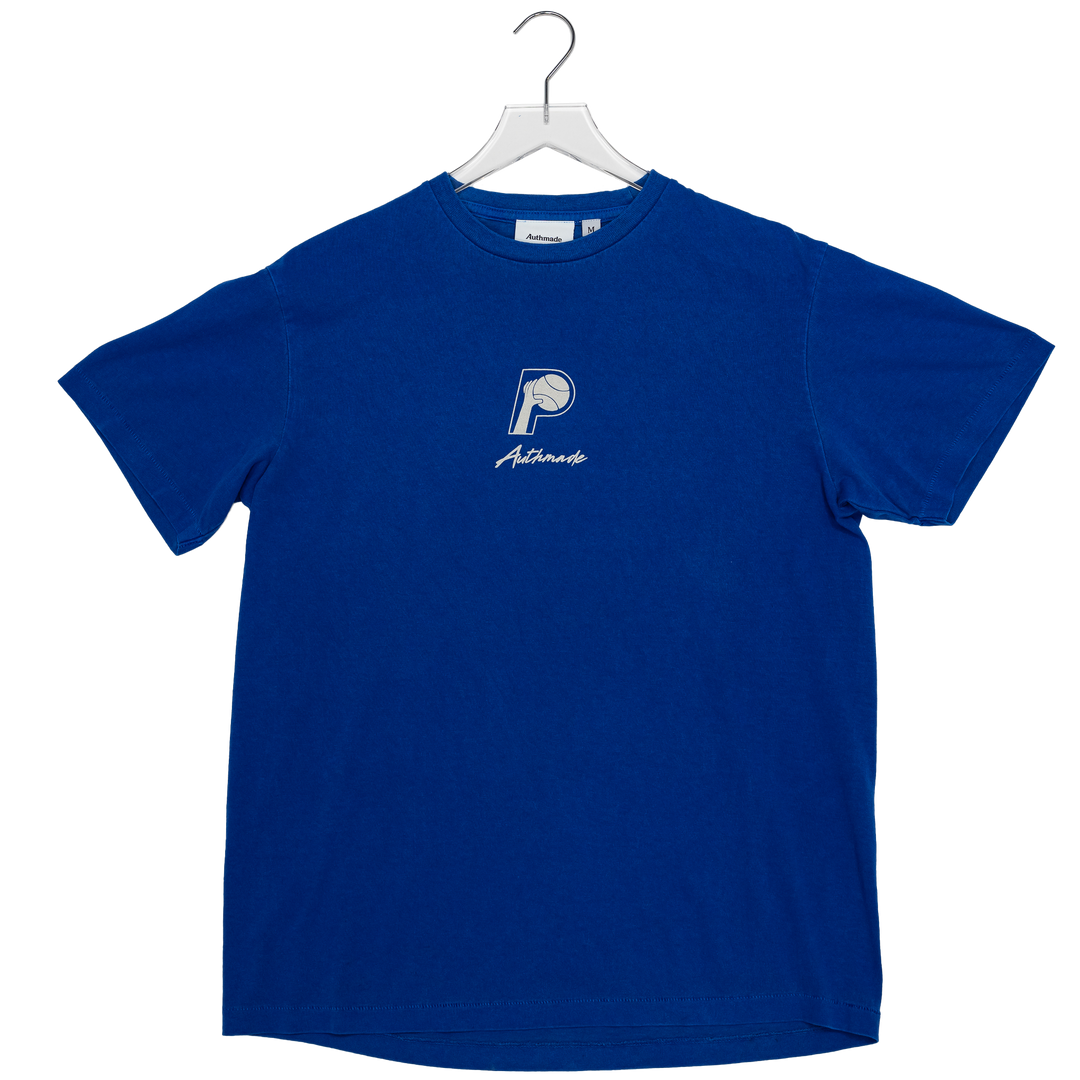 A royal blue short-sleeve t-shirt hanging on a white hanger against a plain white background. The shirt features a small white embroidered logo on the left chest with a basketball graphic and the word &