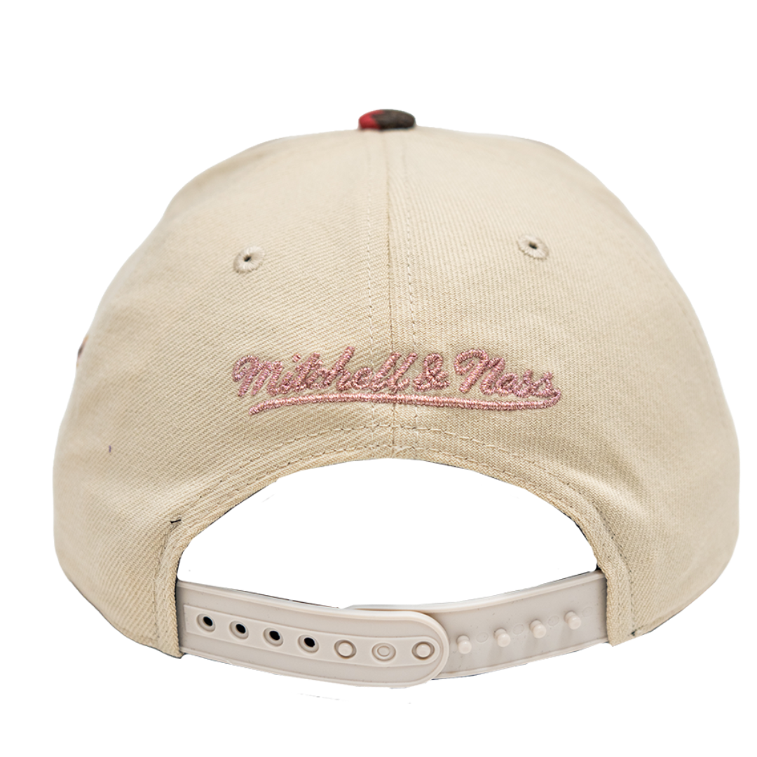 Back view of a beige baseball cap with 'Mitchell & Ness' embroidered in pink script on the front, featuring a beige adjustable strap and plastic snap closure.