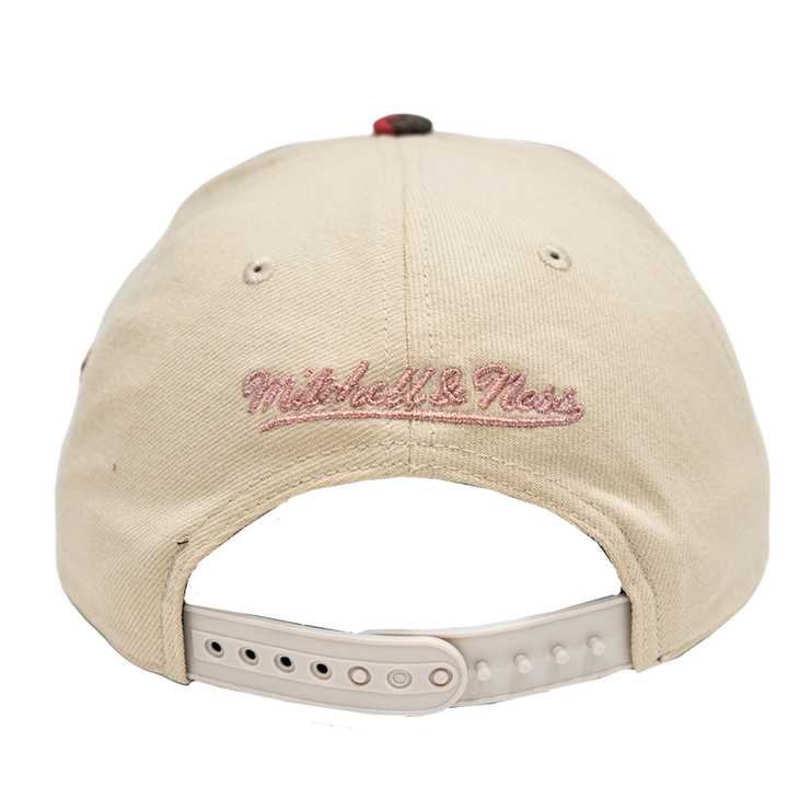 Back view of a beige baseball cap with 'Mitchell & Ness' embroidered in pink script on the front, featuring a beige adjustable strap and plastic snap closure.