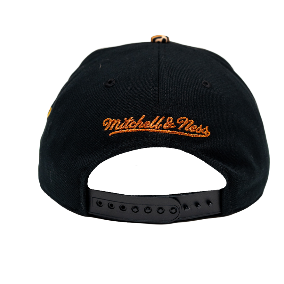 The back of a black Mitchell & Ness baseball cap with orange embroidered script logo and a black adjustable snapback strap.