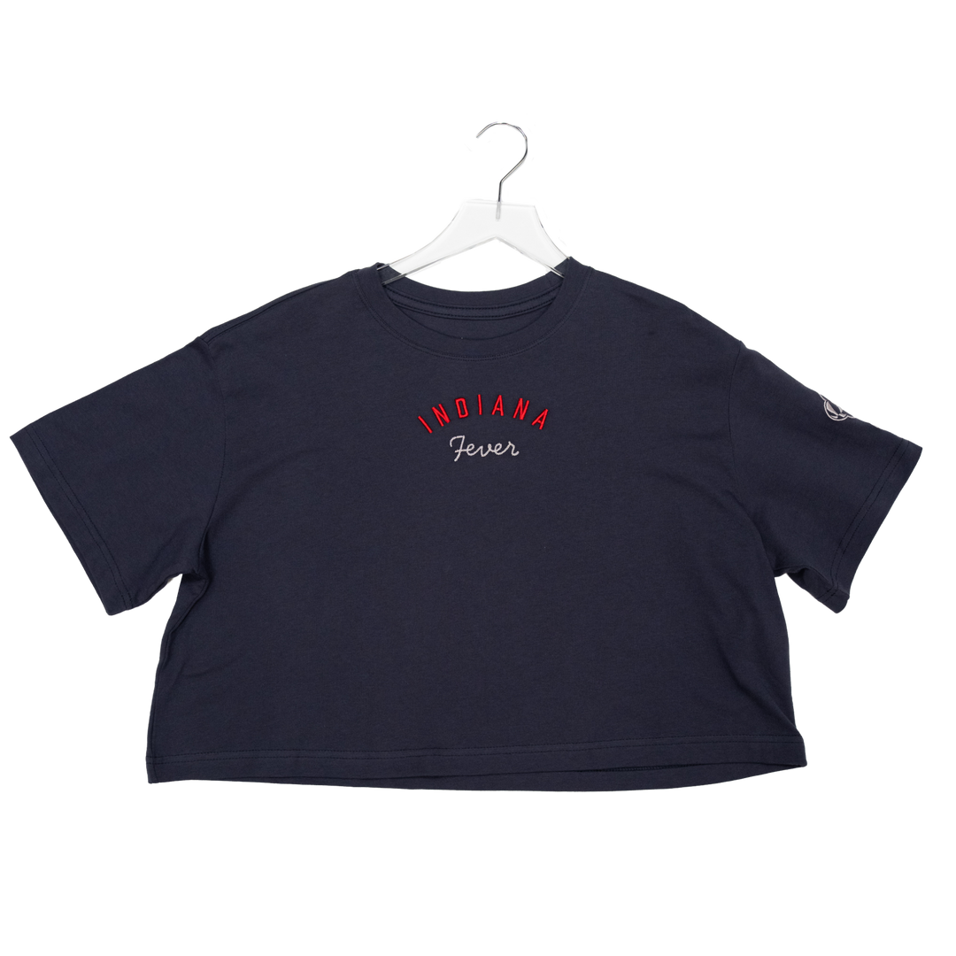 A navy blue short-sleeve crop top t-shirt hanging on a white plastic hanger against a white background. The front features the text 'INDIANA Fever' in red and white script across the chest, and a small white logo on the right sleeve.